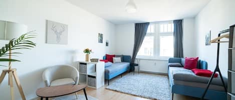 Una sala de estar moderna con un sofá, un sillón y una pequeña mesa. Hay una ventana con cortinas y una planta sobre un atril.