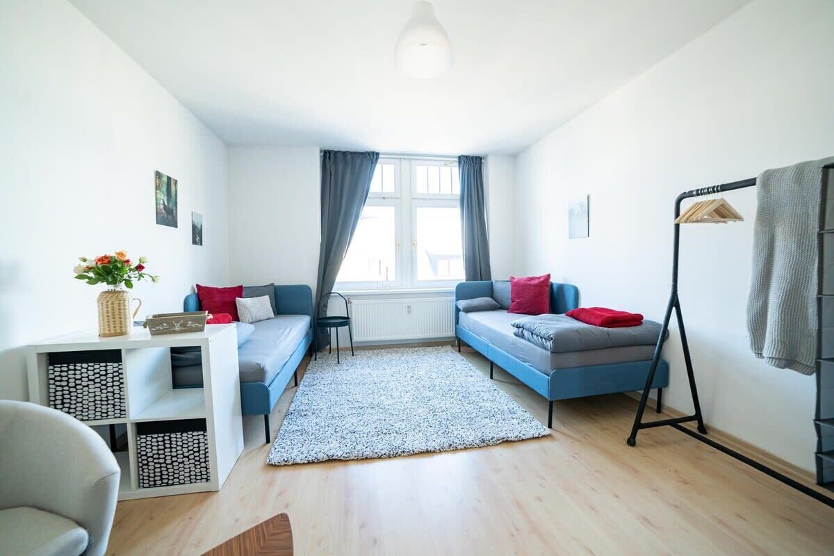 A compact room with a bed, a desk, and a chair. There is a window with curtains, a radiator, and a hanging light fixture.