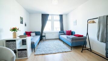 A compact room with a bed, a desk, and a chair. There is a window with curtains, a radiator, and a hanging light fixture.