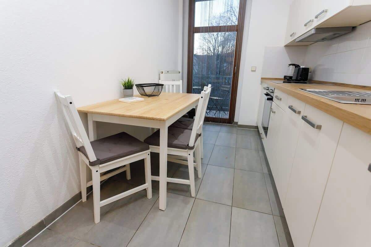 Una piccola cucina con un tavolo di legno e sedie bianche, una finestra con vista sugli alberi e un termosifone bianco.