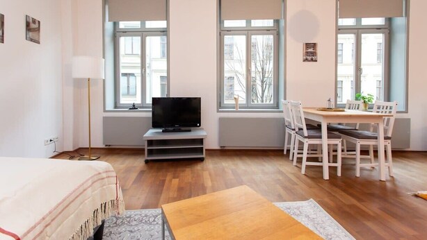 Ein modernes, helles Zimmer mit einem Bett, einem Fernseher auf einem Schrank, einem Esstisch mit Stühlen und großen Fenstern mit Blick auf die Stadt.