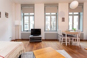 A modern, well-lit room with a bed, a TV on a stand, a dining table with chairs, and large windows offering a view of the city. - FullHouse | Leipzig | M14-1 | PrimeHome (Leipzig)