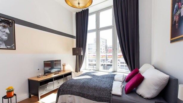 Quarto moderno com cama, escrivaninha, televisão e janela com cortinas.
