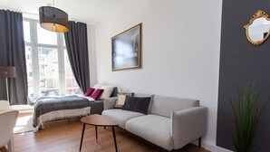 Uma sala de estar moderna com um sofá cinzento, uma mesinha redonda, uma cama com manta cinza e branca, um quadro preto e branco na parede e uma planta em vaso.