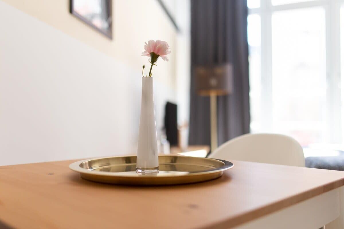 A white vase with a pink flower on a round, gold tray on a wooden table.