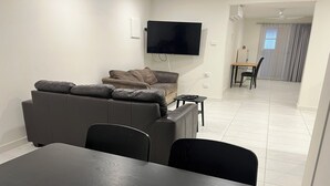 A modern living room with a sofa, a TV mounted on the wall, and a dining area with a table and chairs.