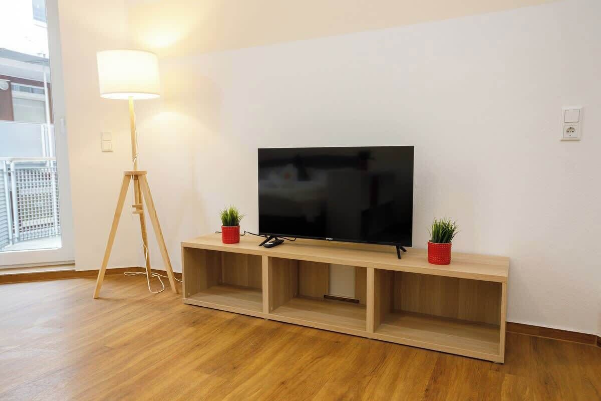 A modern living room with a flat-screen TV on a wooden console, a floor lamp, and two potted plants.