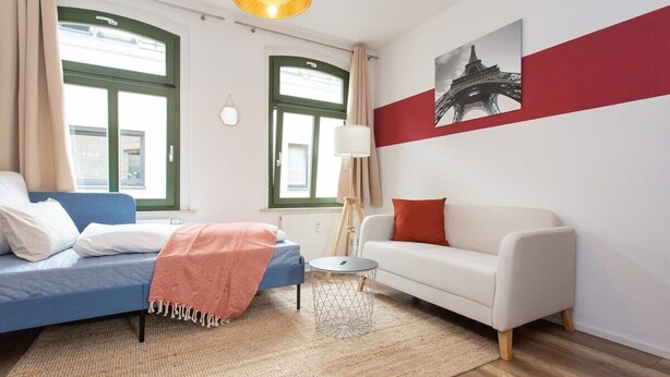 Un salon moderne avec un canapé bleu, un canapé blanc, une petite table ronde et une lampe. Il y a deux fenêtres avec des cadres verts et une bande rouge sur le mur.