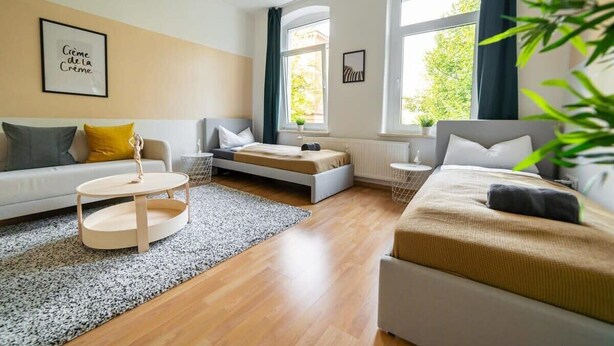 A modern living room with a sofa, a round coffee table, and a bed. There are windows with curtains, a plant, and a framed picture on the wall. - FullHouse | Magdeburg | A9-4 | PrimeApartment (Magdeburg)