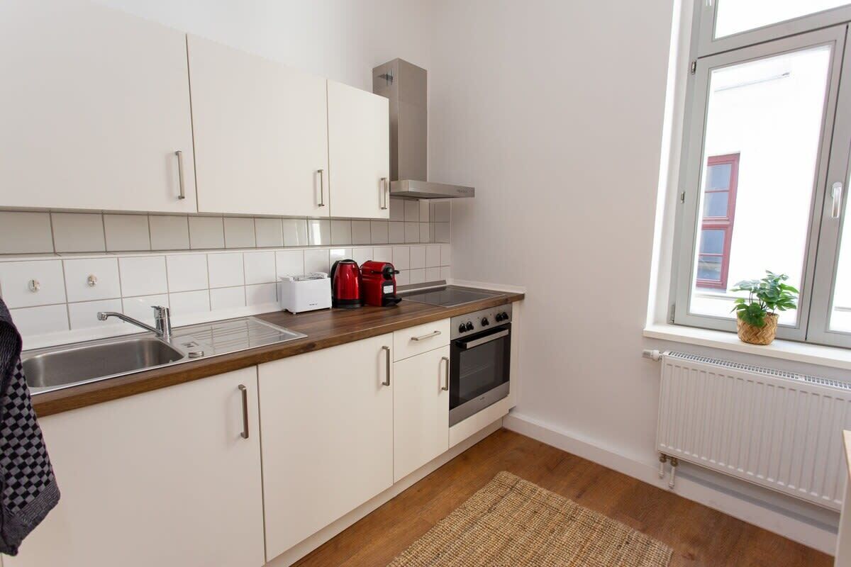 A modern kitchen with white cabinets, a stainless steel sink, a red kettle, and a window with a view of a building outside.