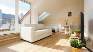 Una sala moderna con un sofá blanco, una mesa de comedor y un televisor. Hay plantas en el suelo y una ventana con vistas a edificios.