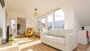 Una sala de estar moderna con un sofá blanco, un sillón amarillo y un televisor sobre un mueble de madera. Hay plantas en macetas y un ventanal grande que proporciona luz natural.