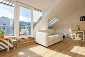 A modern living room with a white sofa, a dining table, and a large window offering a view of the city. - FullHouse Apartments AB5 Rooftop (Halle (Saale))