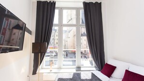 A hotel room with a large window, a bed with red and white pillows, a floor lamp, and a TV mounted on the wall. - FullHouse | Halle | M14 Michelangelo | Zentrum (Halle (Saale))