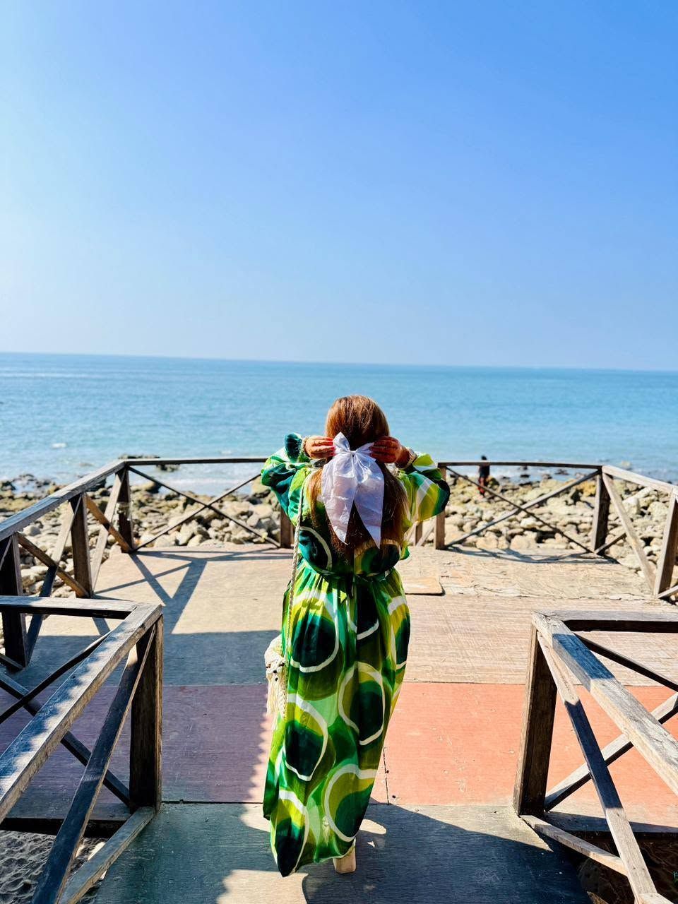 Uma pessoa de vestido verde com laço branco caminhando sobre um calçadão de madeira à beira-mar.