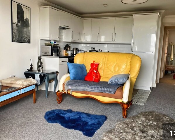 Un espace de vie compact avec un fauteuil jaune, une petite table et un canapé. On aperçoit un coin cuisine au fond.