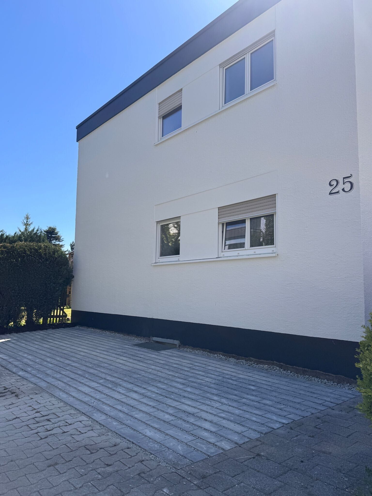 Ein zweistöckiges Gebäude mit weißer Fassade, Hausnummer 25, mit zwei Fenstern im Obergeschoss und einer gepflasterten Auffahrt.
