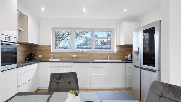 A modern kitchen with white cabinets, a built-in oven, a dining area with a table and chairs, and a window with a view of trees. - Apartment 1 with Private Terrace, Shared Garden, and Wi-Fi (Bad Dürrheim)