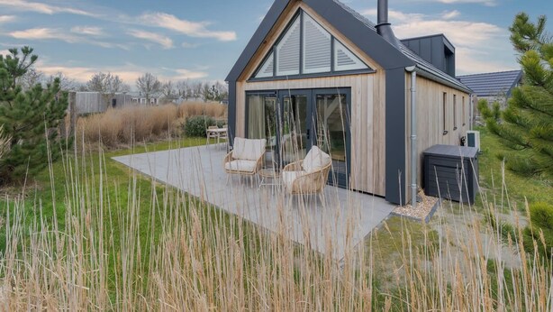 Une maison moderne avec un toit en pente, une cheminée et une terrasse meublée de meubles en osier.