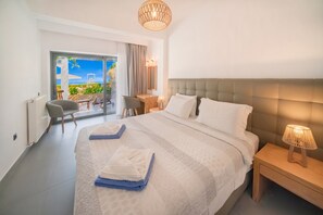 A modern hotel room with a large bed, a bedside table with a lamp, a chair, a small table, and a view of the outdoors. - Barbati Beachfront Pool Villa (Barbati)