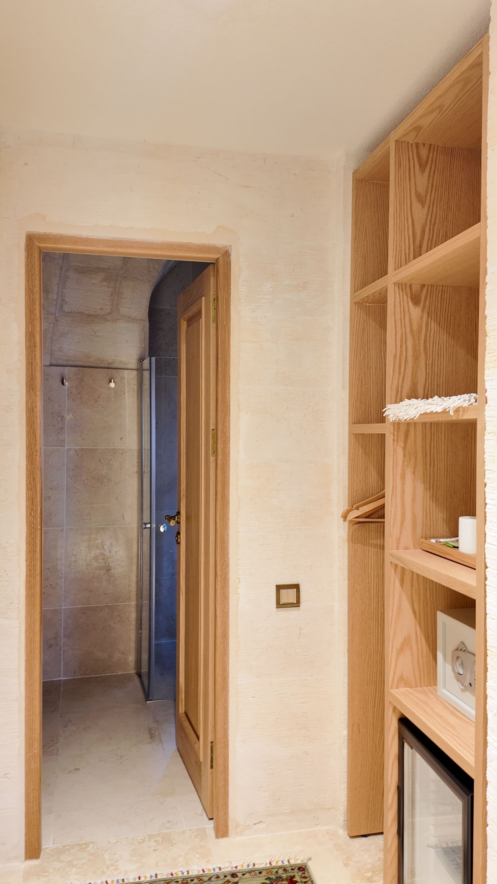 Un corridoio con una porta in legno che dà su un bagno, un mobiletto a mensole in legno con un asciugamano bianco e una piccola presa elettrica sulla parete.