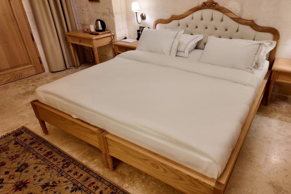 Una cama grande con estructura de madera y ropa de cama blanca, flanqueada por dos mesitas de noche con lámparas, un tablero auxiliar con tetera y una alfombra estampada en el suelo.