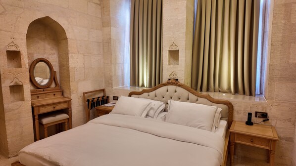 A bedroom with a large bed, wooden nightstands, a vanity with a mirror, and a window with curtains. - Shamas Hotel (Midyat)