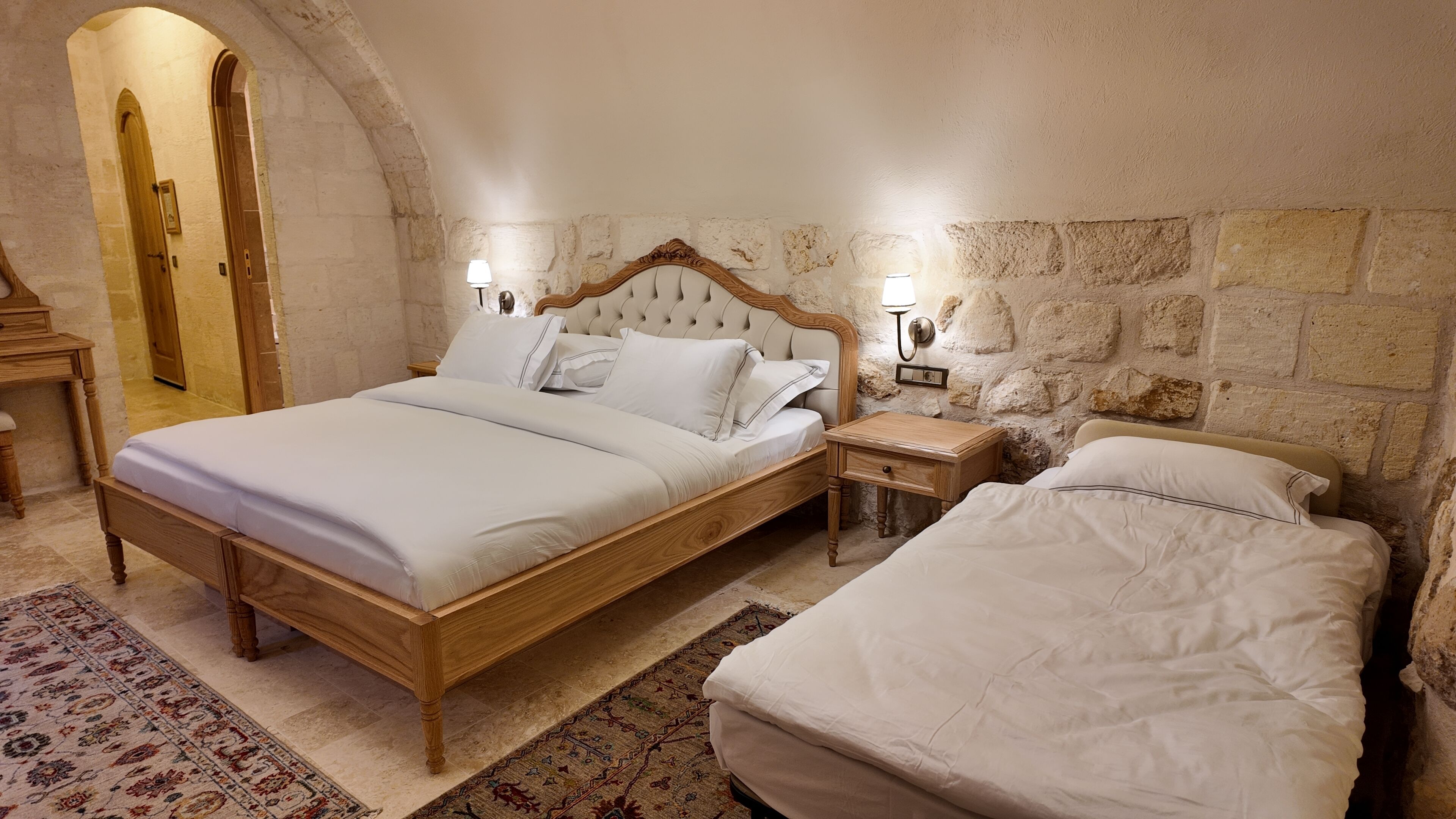 A bedroom with a large bed, a smaller bed, a nightstand, a lamp, and a stone wall.
