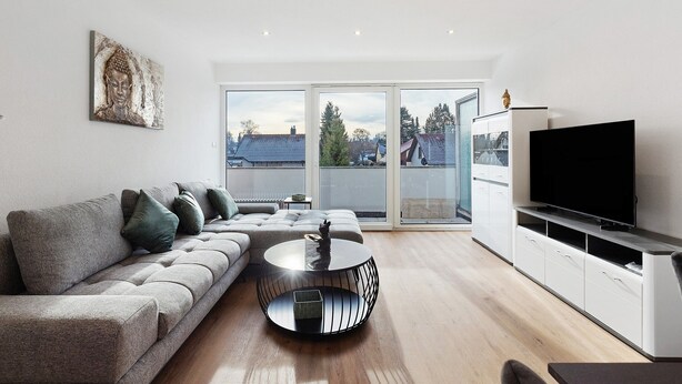 A modern living room with a sectional sofa, a round coffee table, a flat-screen TV, and a large window offering a view of the city. - Apartment '2' with Shared Garden, Balcony, and Wi-Fi (Bad Dürrheim)
