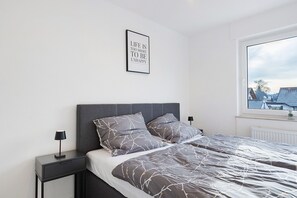 Ein Schlafzimmer mit Bett, Kissen, Nachttisch, Lampe, einem gerahmten Zitat an der Wand und einem Fenster mit Blick.