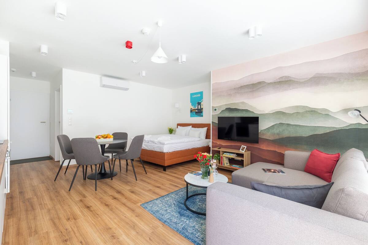 A modern hotel room with a bed, sofa, dining table, and chairs.
