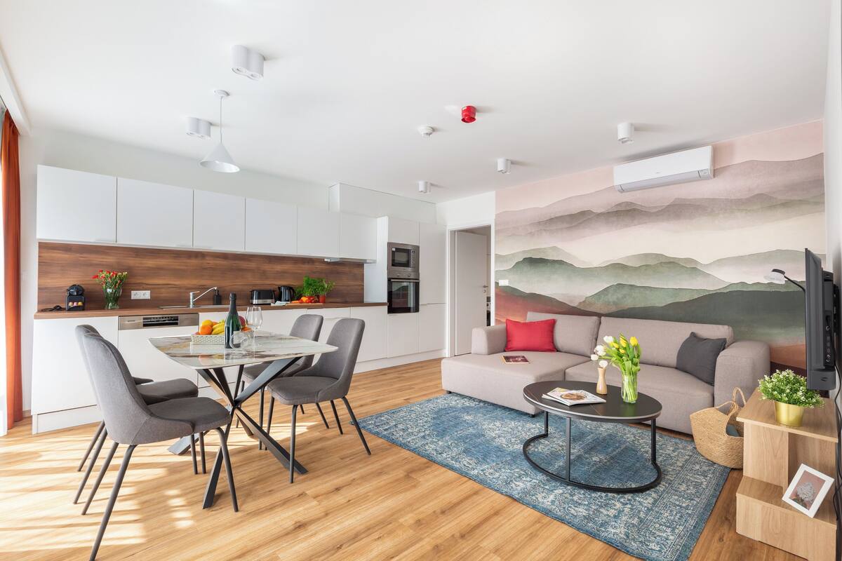 A modern kitchen with white cabinets, a dining area with a glass table and chairs, a living room with a sofa and a TV, and a large wall mural.