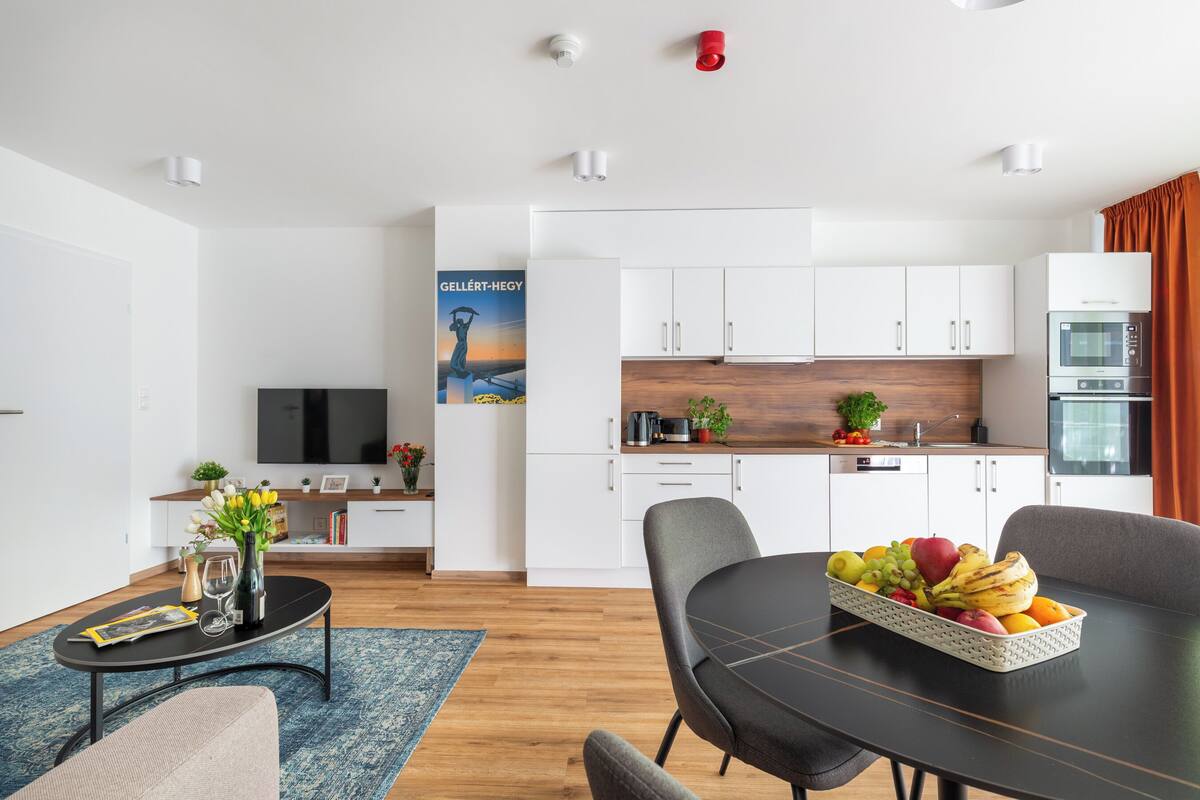 A modern living room with a dining table, a kitchen with white cabinets, and a TV in the background.