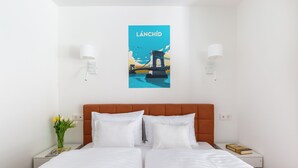 A hotel room with a bed, two bedside tables, a nightstand, a wall-mounted lamp, a poster of a bridge, and a vase with flowers. - BUDAPEST DOWNTOWN RESIDENCE VH (Budapest)