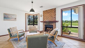 A living room with a fireplace, four chairs, a rug, and a large window with a view of greenery. - Seabreeze Acres ~ Maslin Beach - Seabreeze Acres = Coastal Serenity! (Maslin Beach)