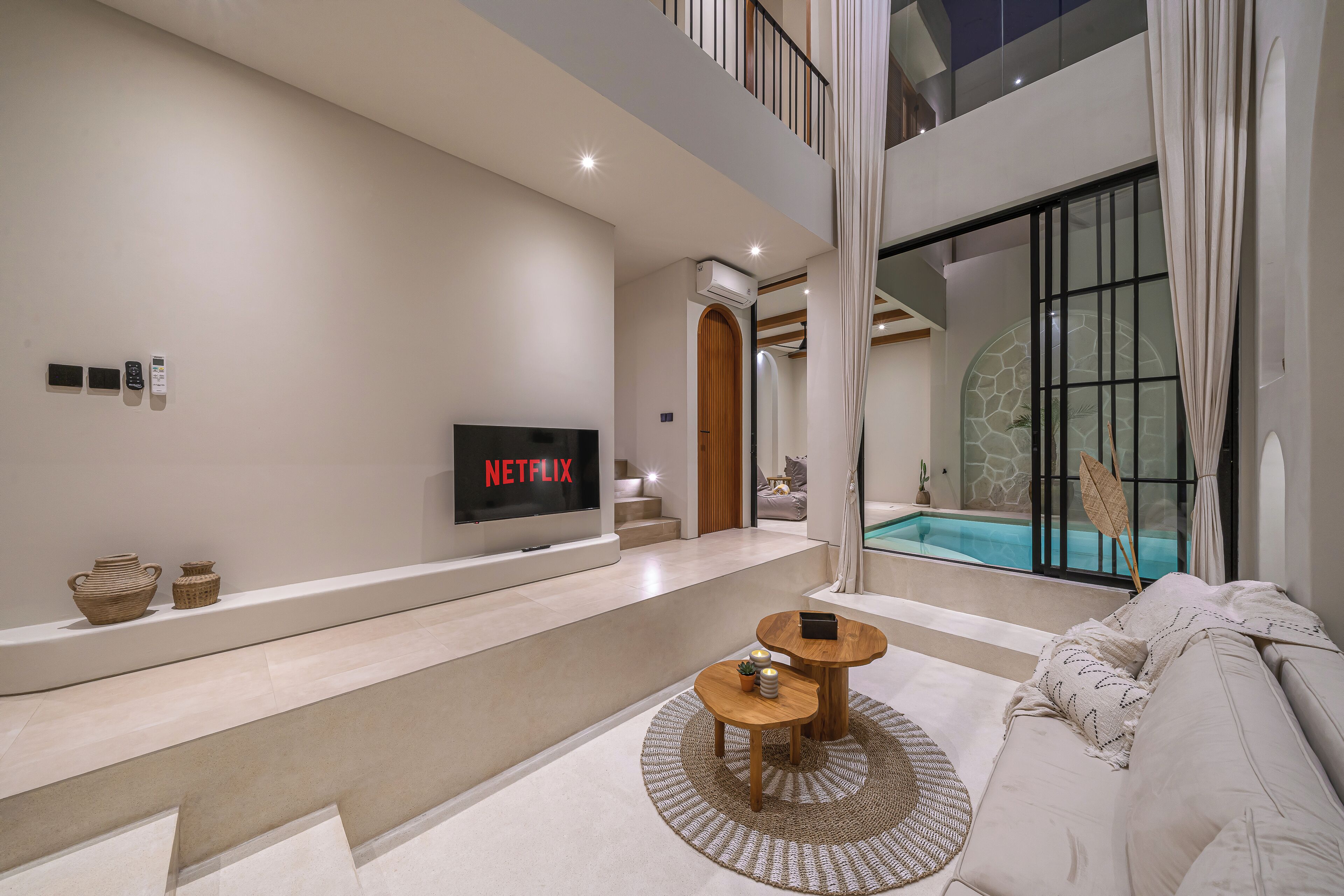 A modern living room with a flat-screen TV, a pool, and a staircase leading to a balcony.