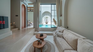 Ett modernt vardagsrum med en beige soffa, ett träbord och en tv på vänster sida. Ett stort fönster ger utsikt över en pool och en palm utanför.
