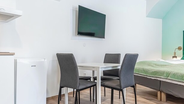 Una habitación moderna con una mesa blanca, cuatro sillas grises, un televisor de pantalla plana montado en la pared, una cama con manta verde y un refrigerador blanco.