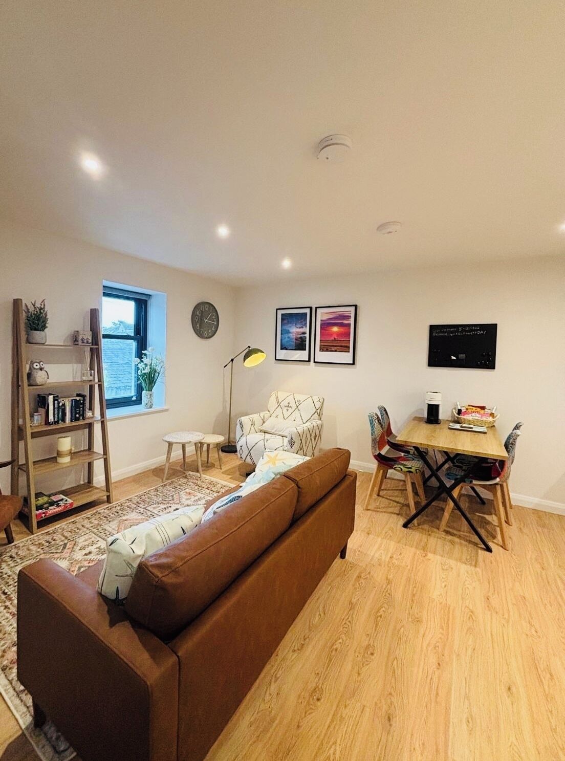 Ein modernes Wohnzimmer mit einem braunen Sofa, Holzboden, einem Esstisch mit Stühlen, einem Bücherregal, einer Stehlampe und einem an der Wand montierten Fernseher.