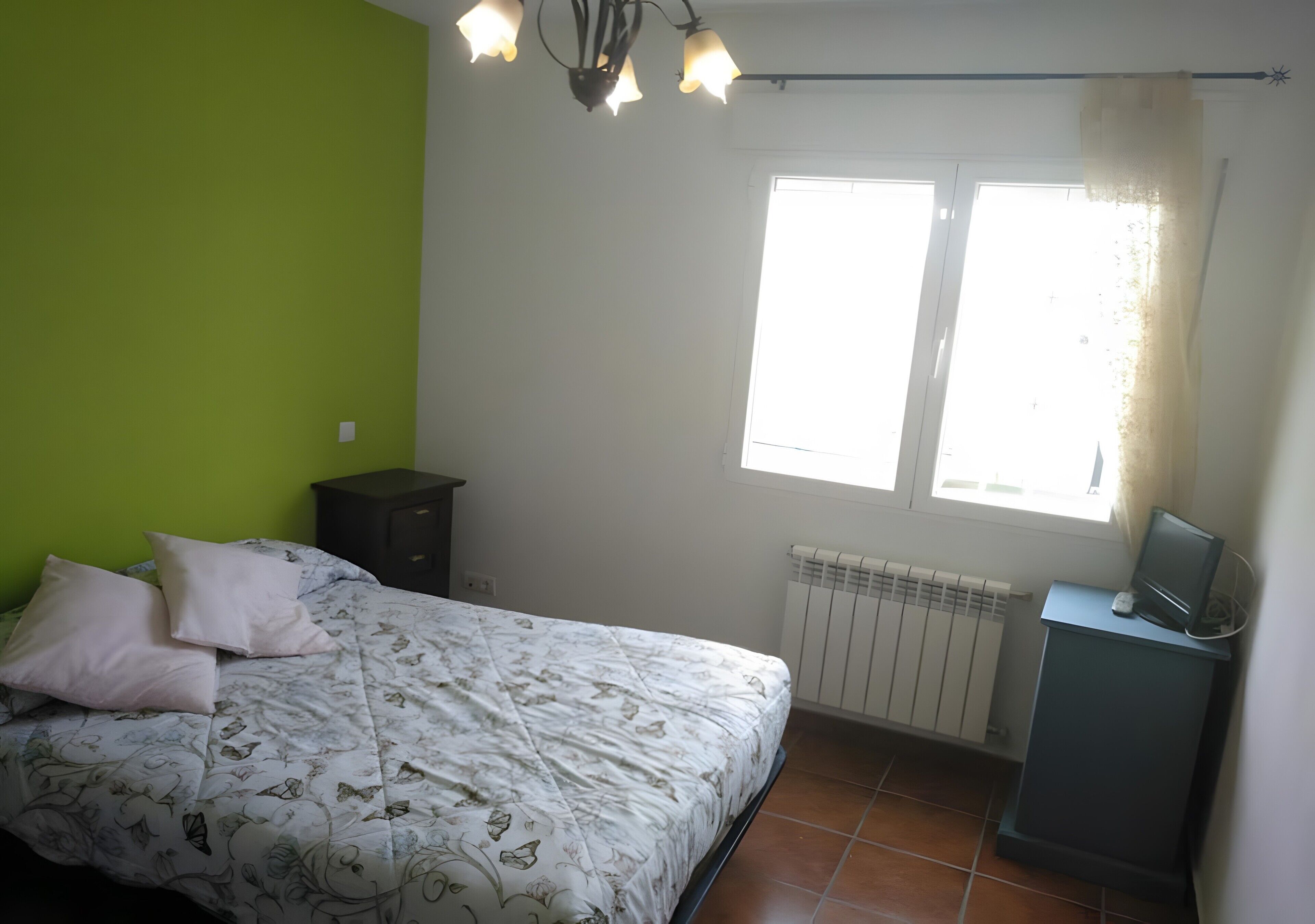 A bedroom with a bed, a nightstand, a desk with a computer, a window with curtains, and a radiator.
