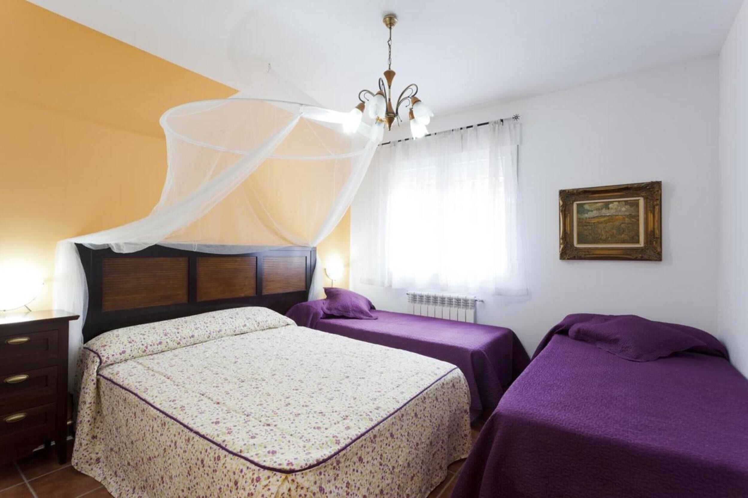 Una camera da letto con un letto, due comodini, un tavolino, una sedia, una finestra con tende leggere, un lampadario e un quadro appeso al muro.