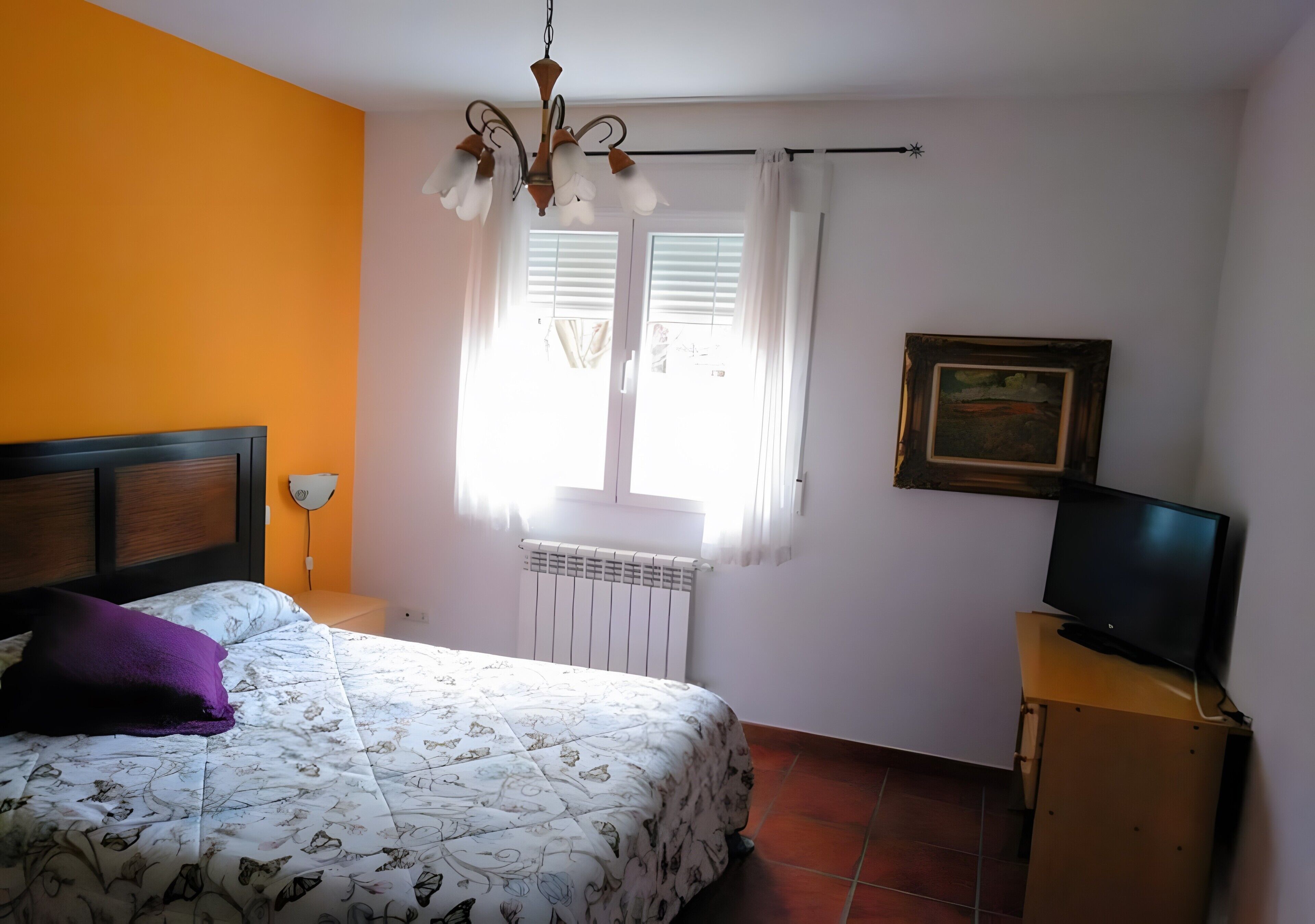 Een slaapkamer met een bed, een televisie op een houten standaard, een raam met gordijnen en een radiator.