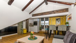 Ein modernes Wohnzimmer mit einem großen Fernseher, einer Essecke mit Stühlen und einer Küche im Hintergrund.