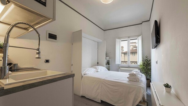 Ett kompakt hotellrum med ett litet pentry, en säng med prydligt hopvikta handdukar, ett fönster med utsikt över byggnader och en platt-TV som är monterad på väggen.