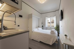 Ett kompakt hotellrum med ett litet pentry, en säng med prydligt hopvikta handdukar, ett fönster med utsikt över byggnader och en platt-TV som är monterad på väggen.