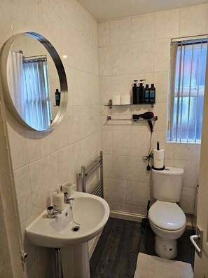 Un baño con espejo redondo, un lavabo, un inodoro, un perchero para toallas y una ventana con cortinas.