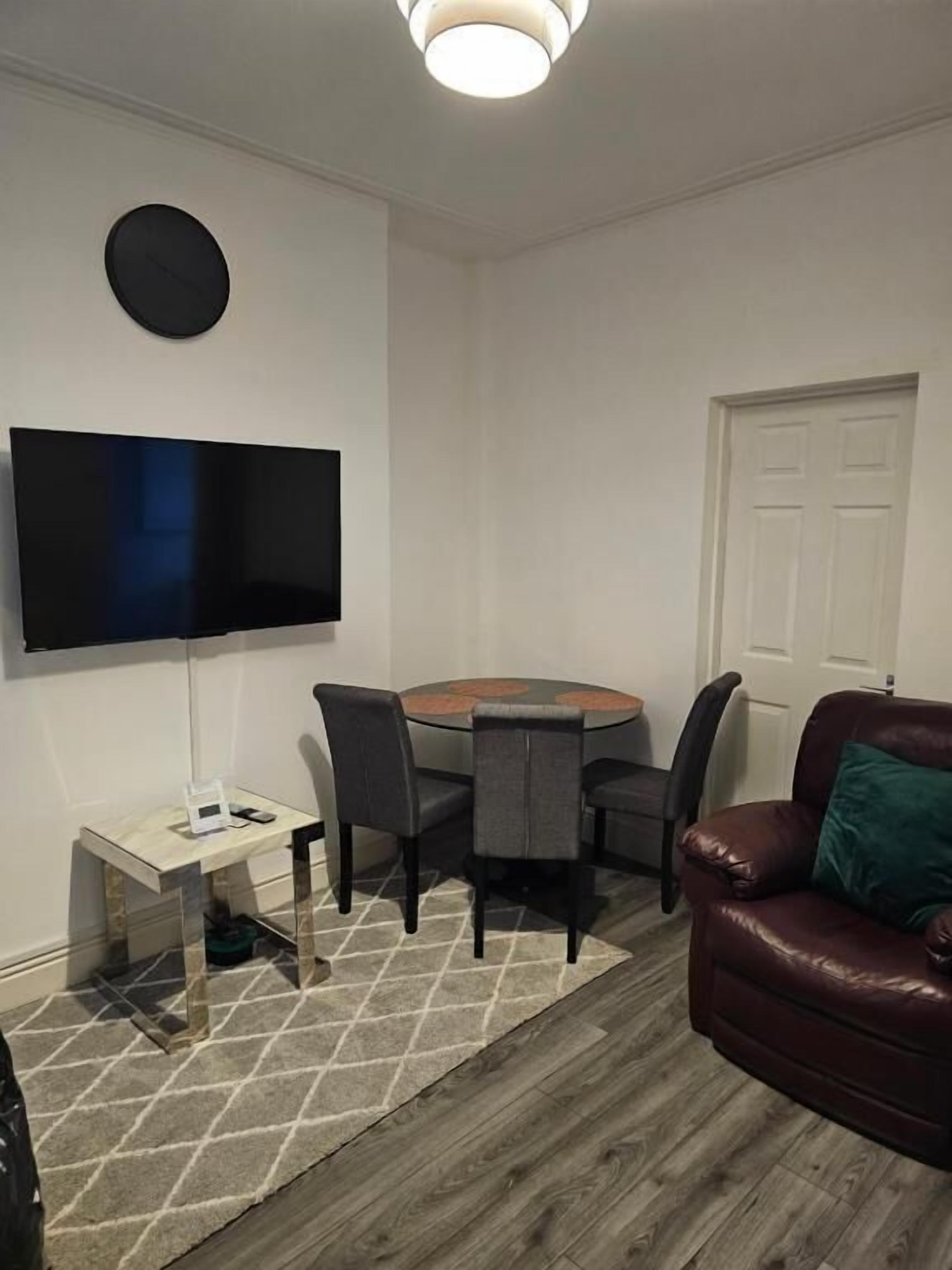 A living room with a round table, four chairs, a TV mounted on the wall, a leather sofa, and a door.
