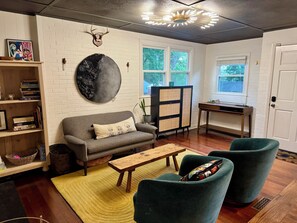 A living room with a sofa, two armchairs, a coffee table, a bookshelf, a decorative wall piece, a cabinet, and a window with a view of trees. - Charming Raleigh Retreat (Raleigh)
