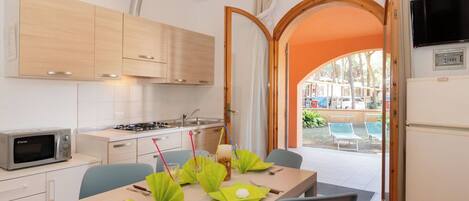 A modern kitchen with wooden cabinets, a microwave, and a stove. There is a dining table set with green napkins and cutlery. An open door leads to an outdoor area with a pool and lounge chairs.