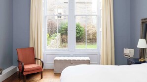 A room with a large window, a chair, a bedside table with a lamp, and a bed.
